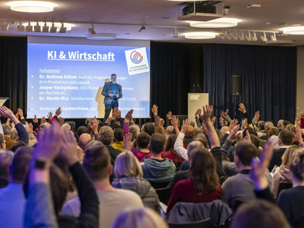 Viele Besucherinnen und Besucher sitzen bei einer Veranstaltung der Heilbronner KI-Gespräche in einem Vortragssaal und melden sich während einer Diskussion zu Wort. Auf der Bühne ist eine Präsentation zum Thema „KI & Wirtschaft“ zu sehen.