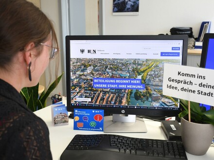 Eine Frau sitzt an einem Schreibtisch und betrachtet auf einem Computerbildschirm die Website der Heilbronner Bürgerbeteiligung. Neben dem Monitor steht ein Schild mit der Aufschrift „Komm ins Gespräch – deine Idee, deine Stadt!“.