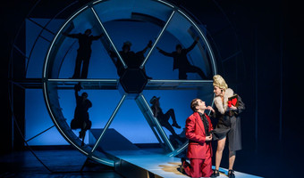 Das Bild zeigt eine Schauspielerin und einen Schauspieler vor einem Bühnenbild mit Schattenelementen.