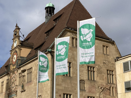 Das Bild zeigt drei wehende grüne Farben mit der weißen Friedenstaube vor dem Rathaus. Es sind Flaggen der internationalen Organisation "Mayors for Peace", übersetzt Bürgermeister für Frieden.